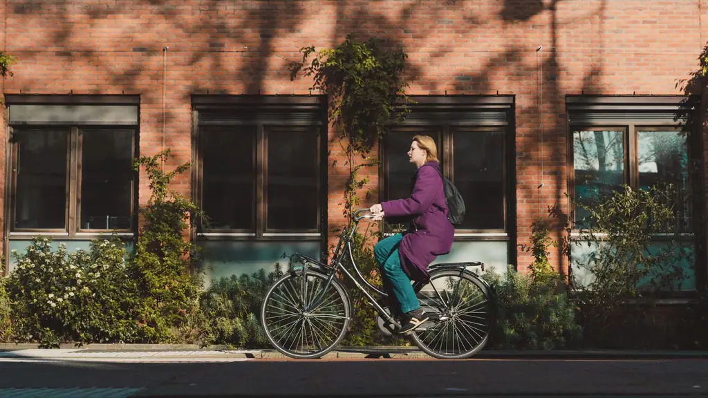 Vrouw in een paarse jas fietst langs een bakstenen gevel, boomschaduwen op de muur
