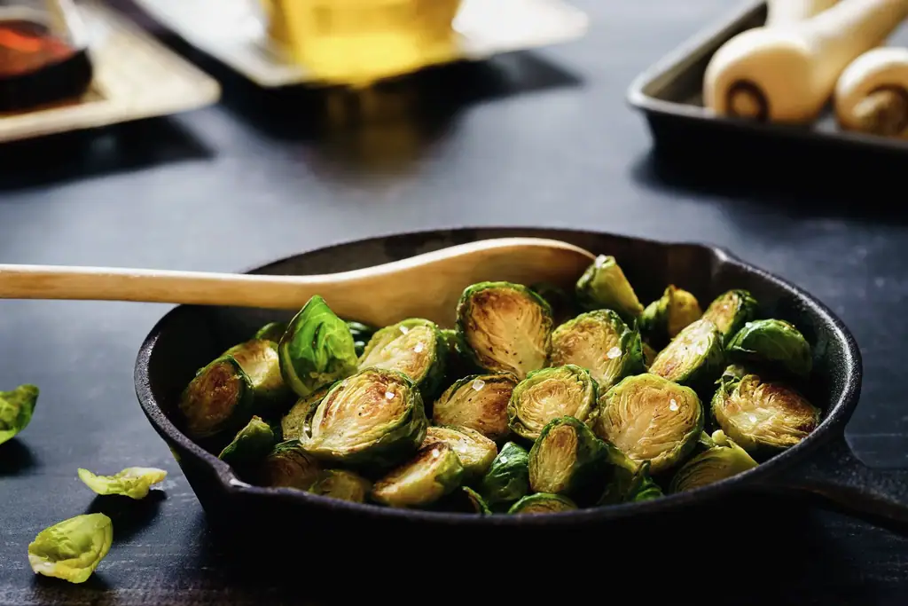 Gietijzeren pan met gebakken spruitjes en een houten lepel op een donkere tafel, warm licht