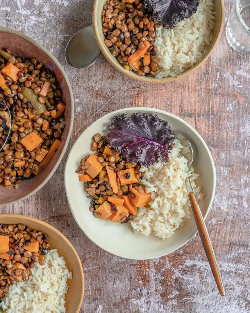 Van bovenaf: kommen linzenstoof met zoete aardappel, rijst en boerenkool op een houten tafel, een glas water